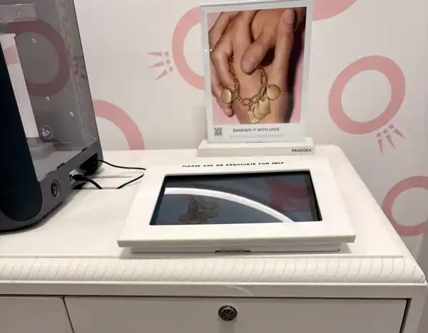 Tablet kiosk in a white acrylic enclosure on the Pandora engraving station console, next to the engraver